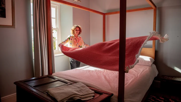 Una persona está junto a una cama con dosel, haciendo la cama con una colcha roja sobre sábanas blancas.
