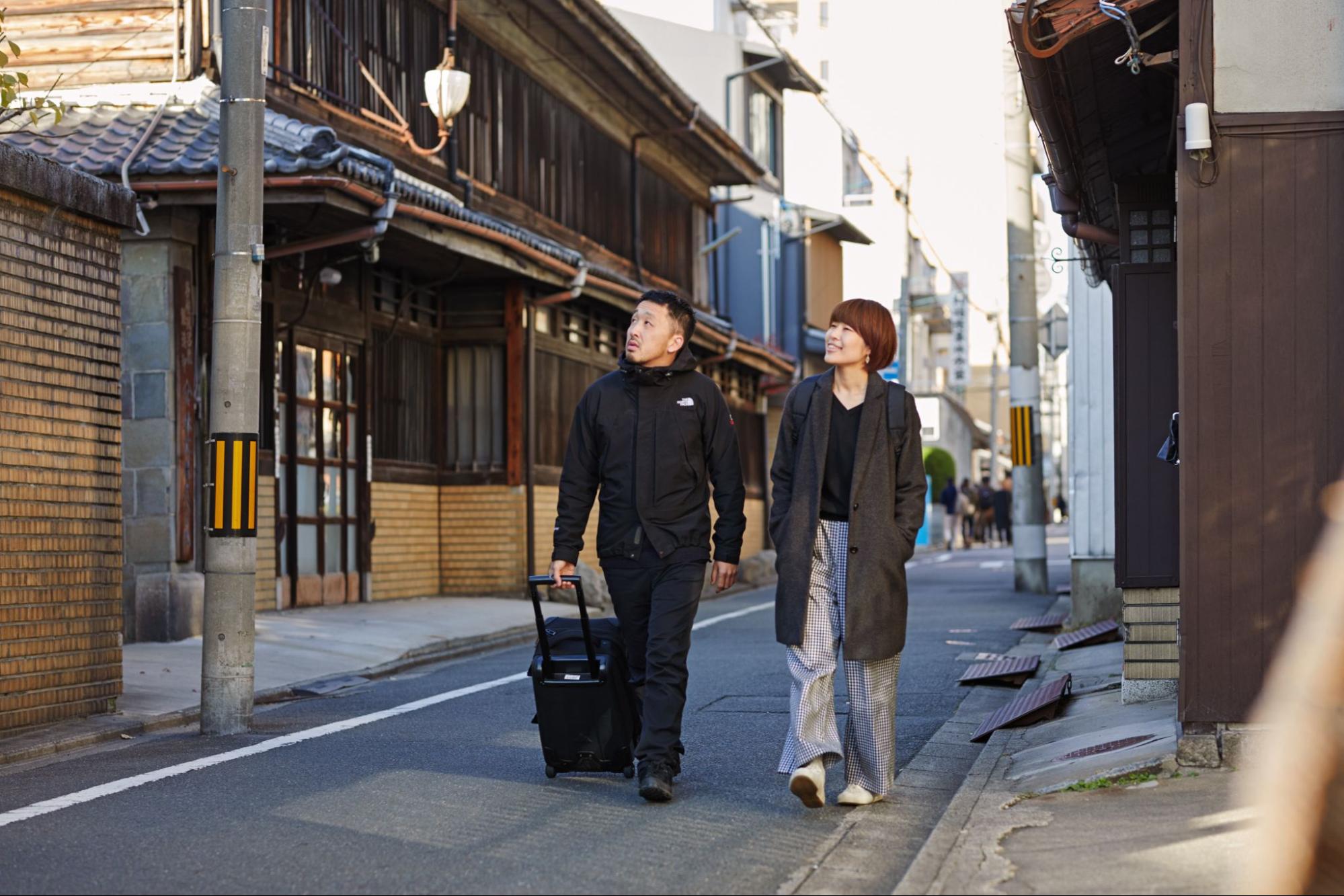 Երկու Airbnb հյուր․ մեկը քաշում է անիվներով ճամպրուկ։ Նրանք քայլում են Տոկիոյի փողոցներից մեկով և նայում են վեր՝ աղյուսե շենքի։