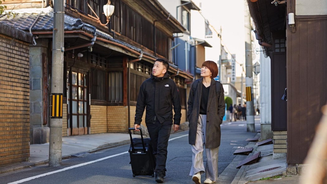 Zwei Gäste auf Airbnb, von denen eine Person einen Rollkoffer zieht, gehen durch eine Straße in Tokyo. Sie haben die Köpfe in Richtung eines Backsteingebäudes gedreht.