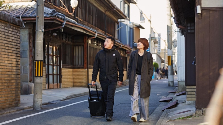 Dos huéspedes en Airbnb levantan la vista hacia un edificio de ladrillos mientras caminan por una calle de Tokio. Uno de ellos jala una maleta con ruedas.