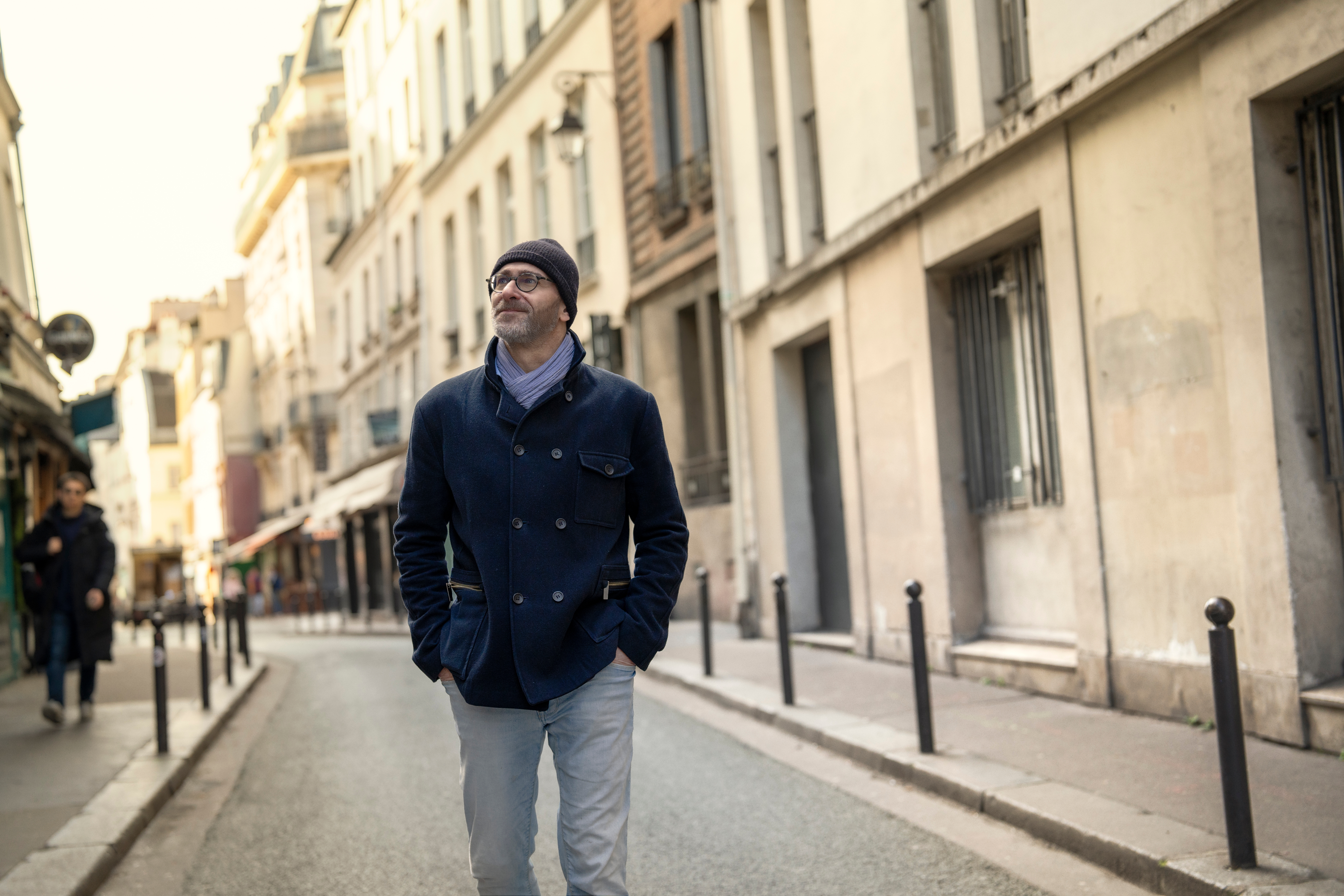 Una persona amb ulleres i una jaqueta blava camina per un carreró empedrat a París.