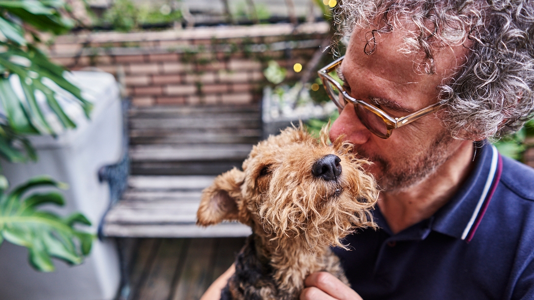 A person wearing glasses and a polo shirt kisses and pets a dog on an outdoor wood deck with a garden bench.