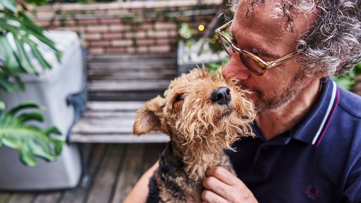Osoba sa naočarima, u polo majici, ljubi i mazi psa na spoljnoj drvenoj terasi sa baštenskom klupom.