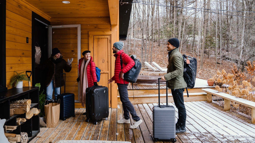 Cuatro huéspedes con sombreros y abrigos de invierno cargados con mochilas y maletas de ruedas caminan sobre una terraza de madera para entrar en un alojamiento.