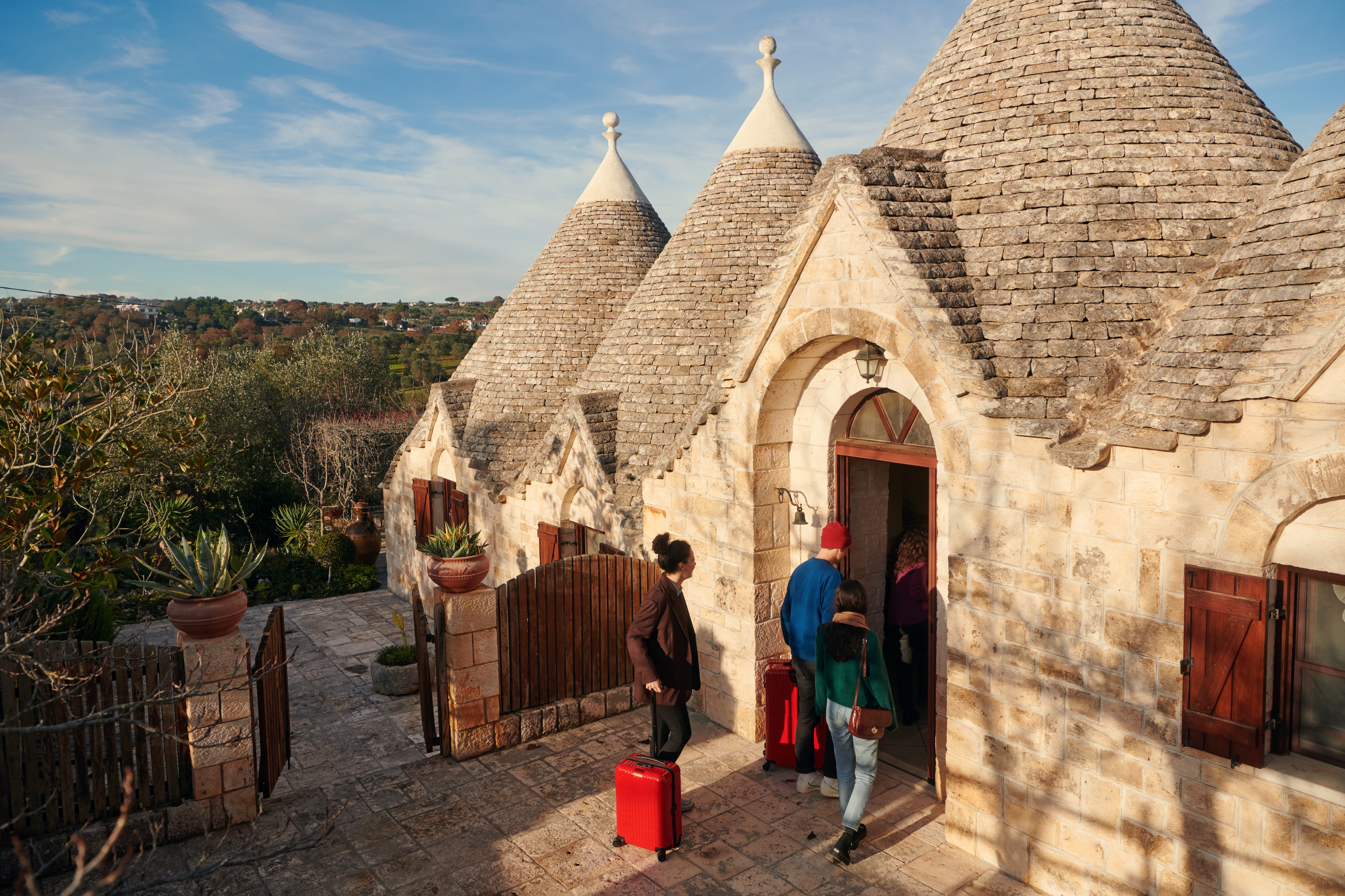 Drei Personen, die zwei rote Koffer ziehen, gehen durch eine Tür in ein Steinhaus, von dem aus man auf Bäume und Dächer blickt.
