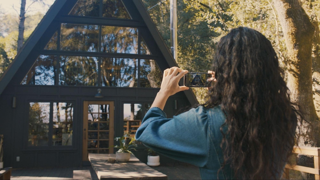 A person with long dark hair is photographing an A-frame cabin in the woods with a mobile phone.