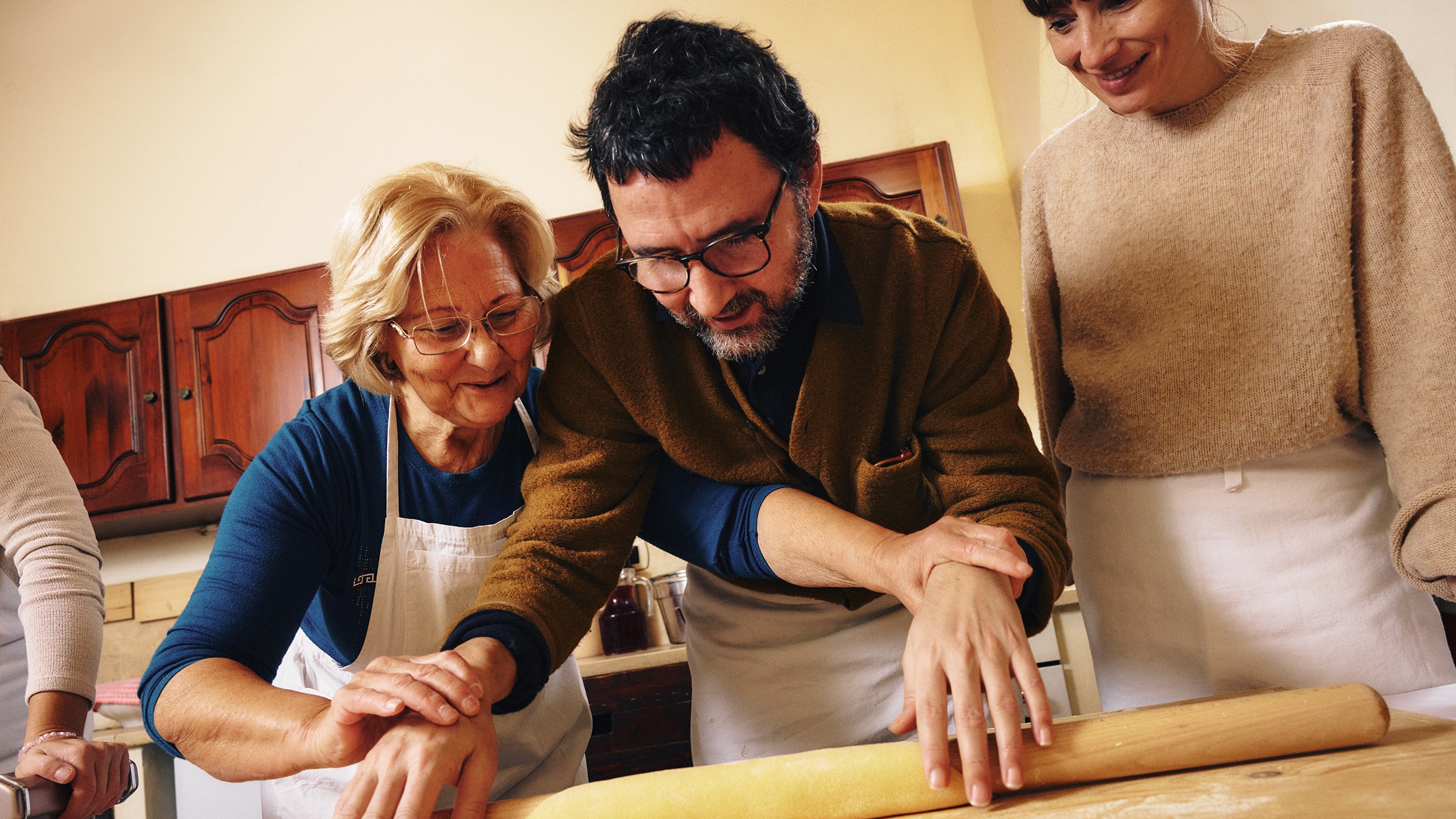 Човек с бяла престилка се усмихва, докато направлява ръцете на друг човек, който разточва тесто с точилка.
