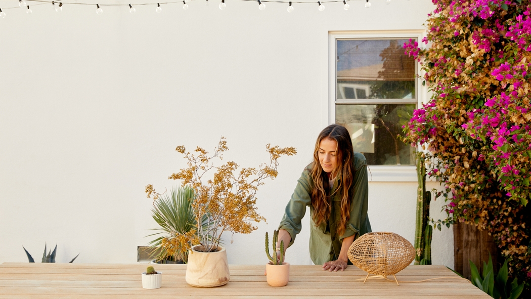 A person adjusts a cactus in a pink pot on top of a wooden table with a potted plant in the foreground.