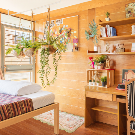 Zonlicht overspoelt een slaapkamer met een smal bureau en planken die in houten lambrisering zijn ingebouwd. Een tiental planten zorgt voor een natuurlijke sfeer.
