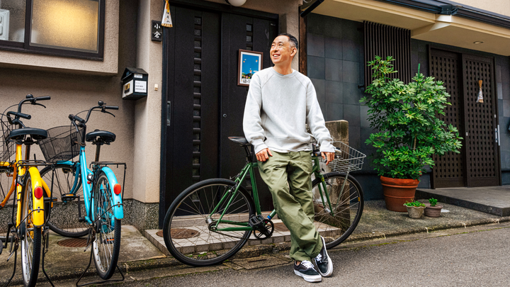 Una persona con una felpa grigia e dei pantaloni verdi posa appoggiata a una bicicletta davanti a una porta nera e a delle piante in vaso.