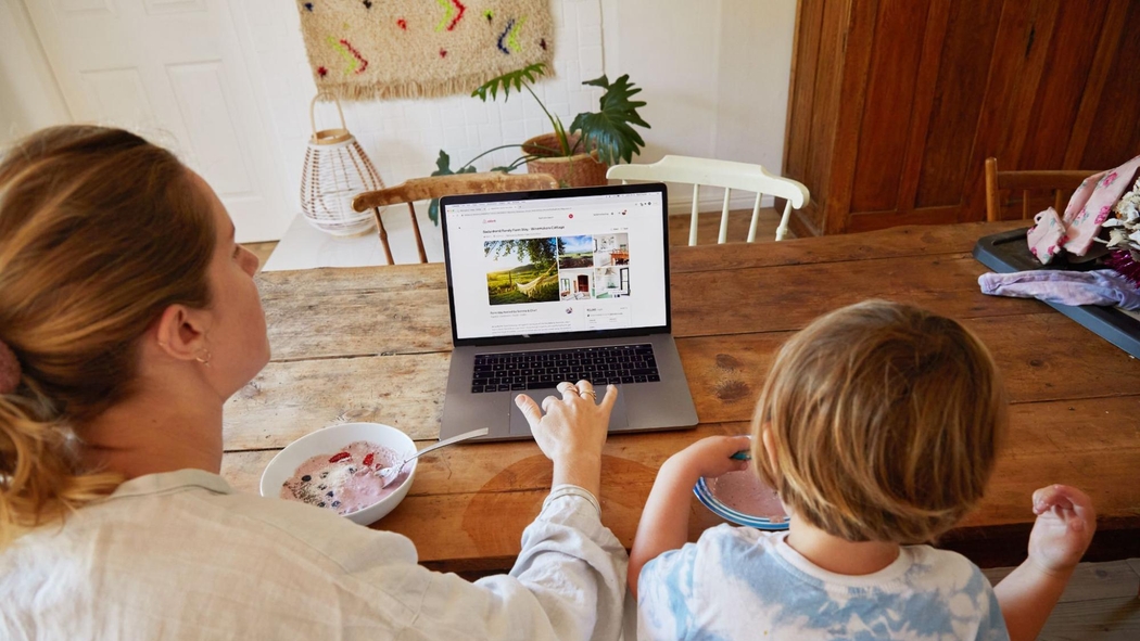 Un adulto revisa un anuncio en Airbnb en una laptop mientras está sentado en una mesa de madera con un niño pequeño. Ambos están comiendo yogur y fruta.