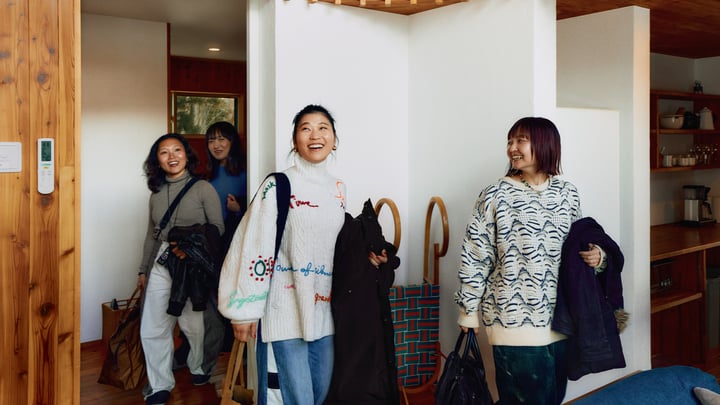 Cuatro personas sonrientes que llevan en la mano chaquetas, bolsas de tela y más equipaje entran en un alojamiento.