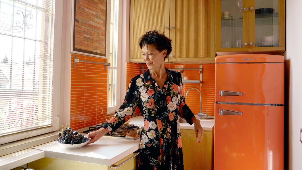 A person places a bowl of grapes on a kitchen surface, with an orange fridge and orange tiles in the background.
