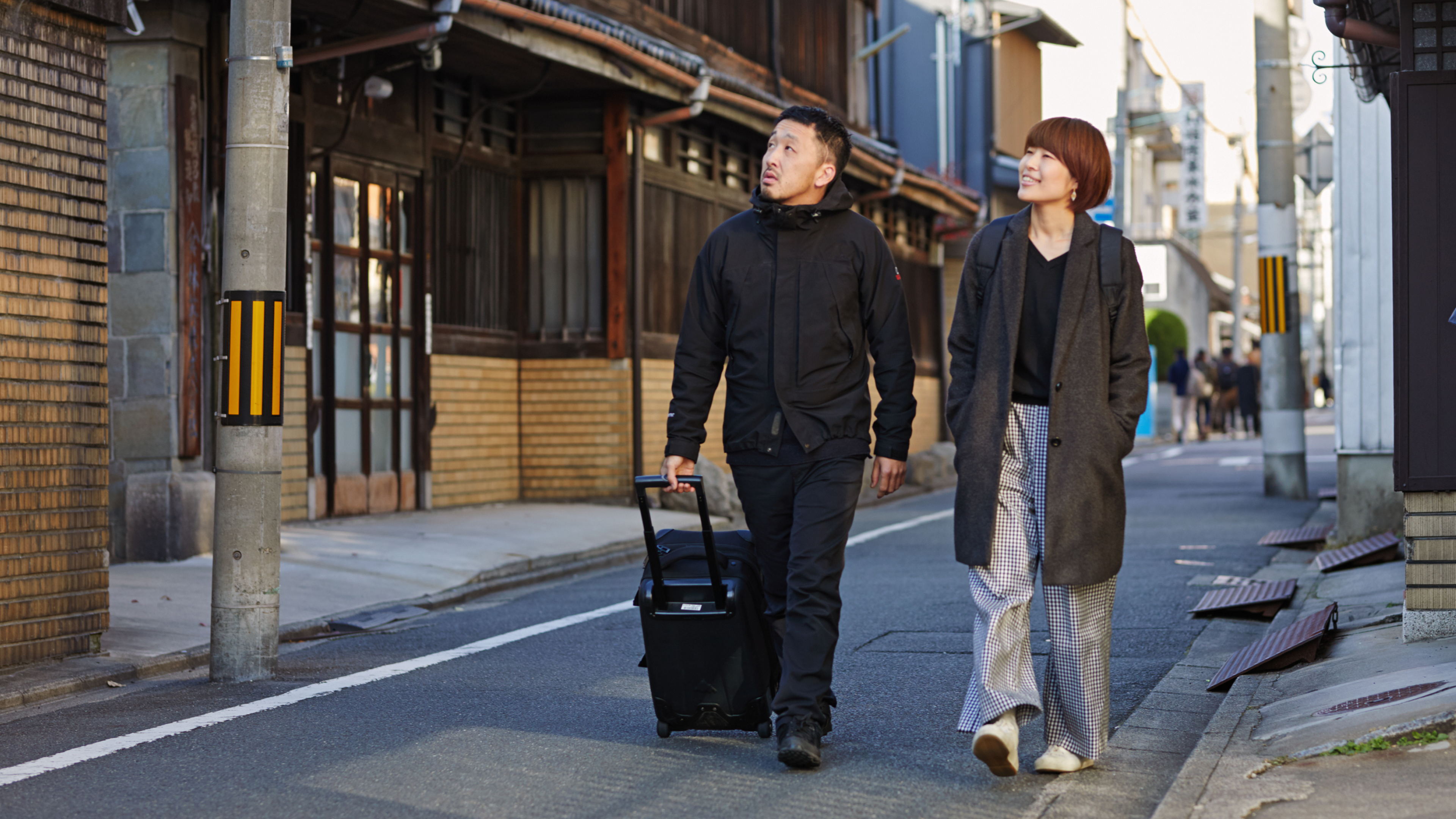 Two Airbnb guests, one pulling a wheelie bag, look up at a brick building as they walk down a street in Tokyo.