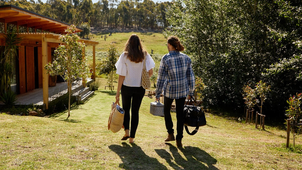 On a sunny day, two people walk down a grassy hill to a modern cabin with a porch and many windows, luggage in their hands.