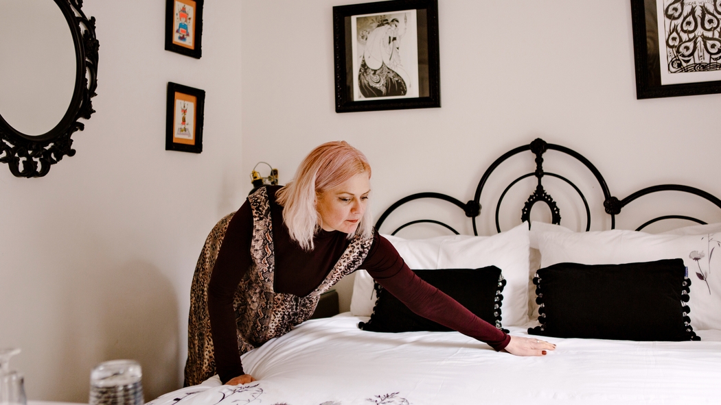 En person i en kjole med leopardprint og en langærmet bordeauxfarvet skjorte udglatter tæppet på en seng i sort-hvid.