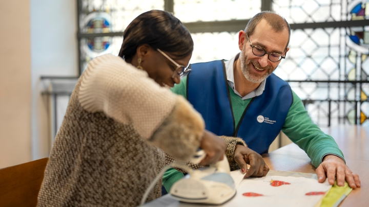 En sittande person stryker ett tygstycke på ett bord medan en annan sittande person tittar på.