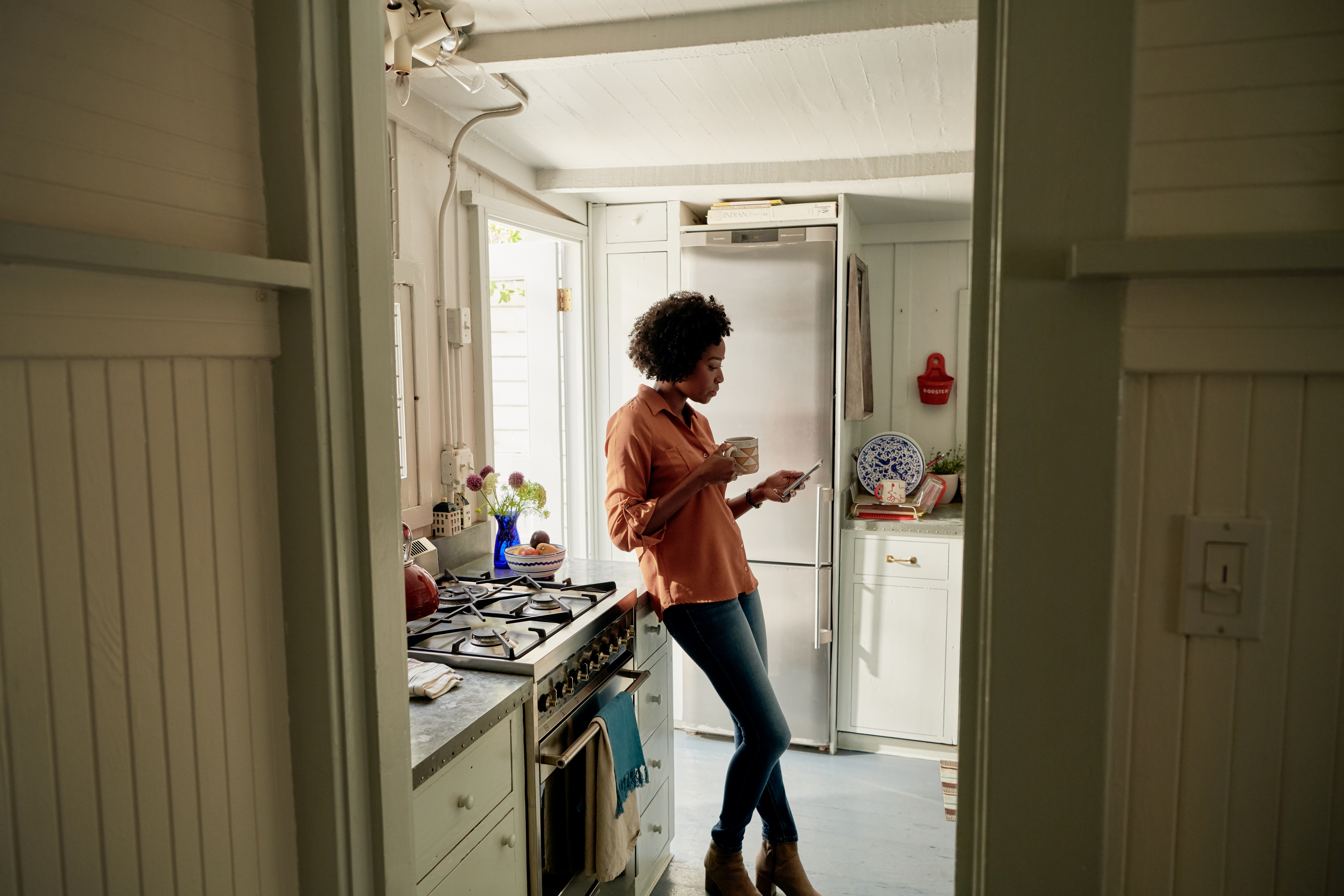 Una persona che indossa dei jeans e una camicia arancione è appoggiata al bancone di una cucina accanto ai fornelli a gas, con in mano una tazza e uno smartphone.