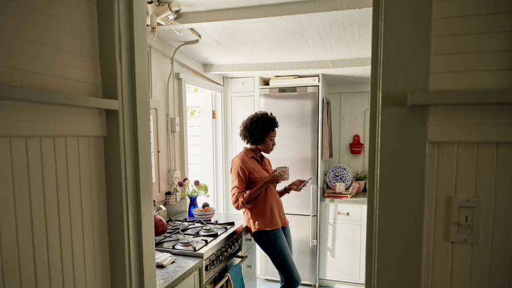一个穿着牛仔裤和橙色衬衫的人拿着杯子和智能手机，倚在灶台边的厨房操作台上。