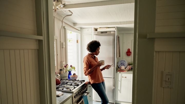 Une personne portant des jeans et une chemise orange s'appuie sur un comptoir de cuisine à côté d'une cuisinière à gaz et tient dans ses mains une tasse et un téléphone intelligent.