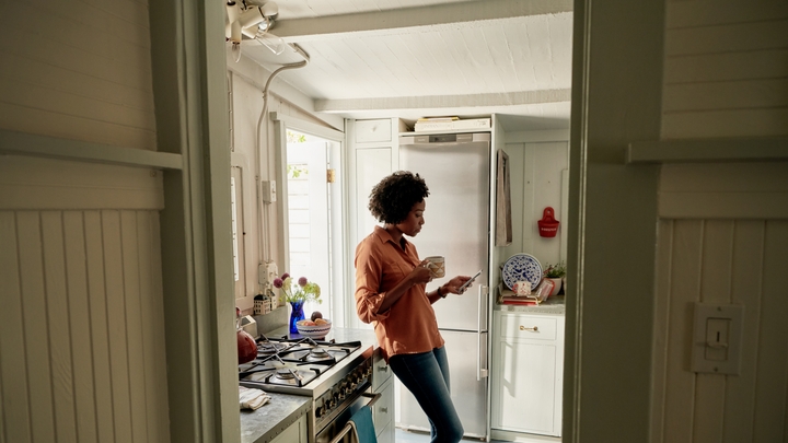 A person in jeans and an orange shirt leans on a kitchen counter next to a gas range, holding a mug and a smartphone.