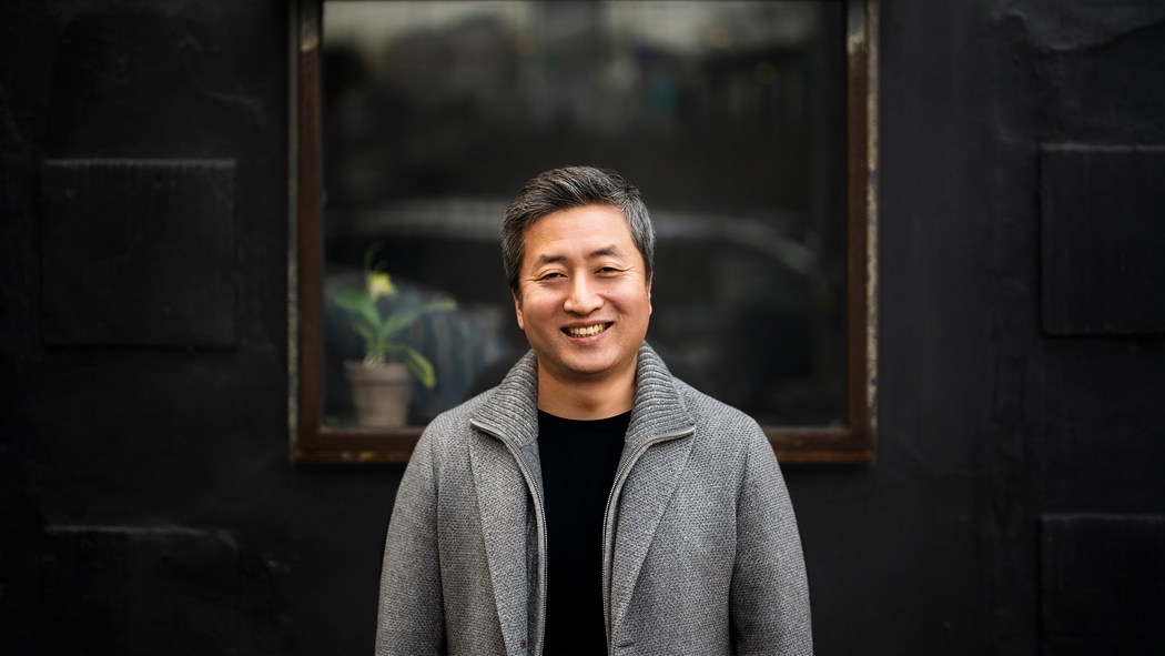En smilende mand iført en grå sweaterblazer står foran et vindue mod en mørk væg.