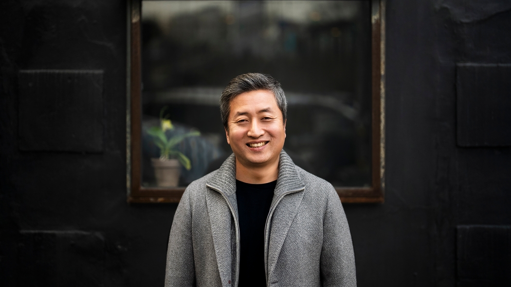 Un hombre sonriente que lleva una chaqueta de suéter gris está de pie frente a una ventana contra una pared oscura.