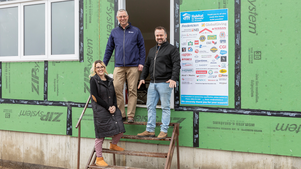 Three individuals stand on metal steps in front of a duplex under construction by Habitat for Humanity New Brunswick.