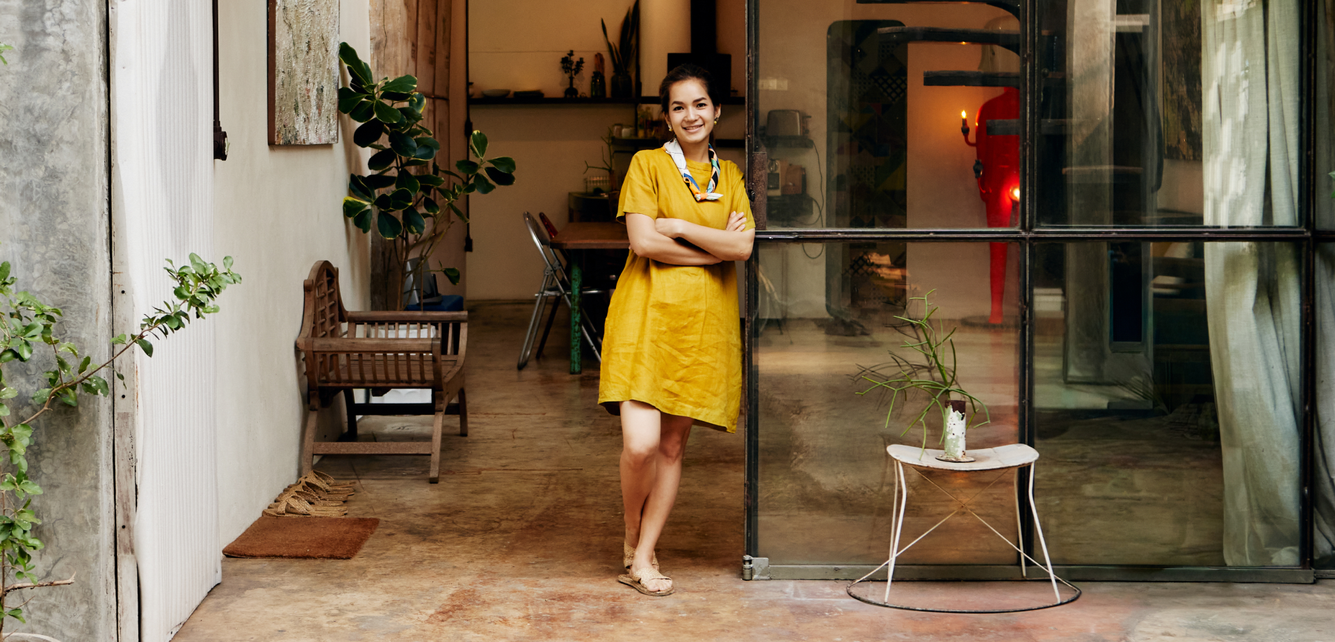A person leans against a floor-to-ceiling window with a small table beside them and a larger table behind them. 