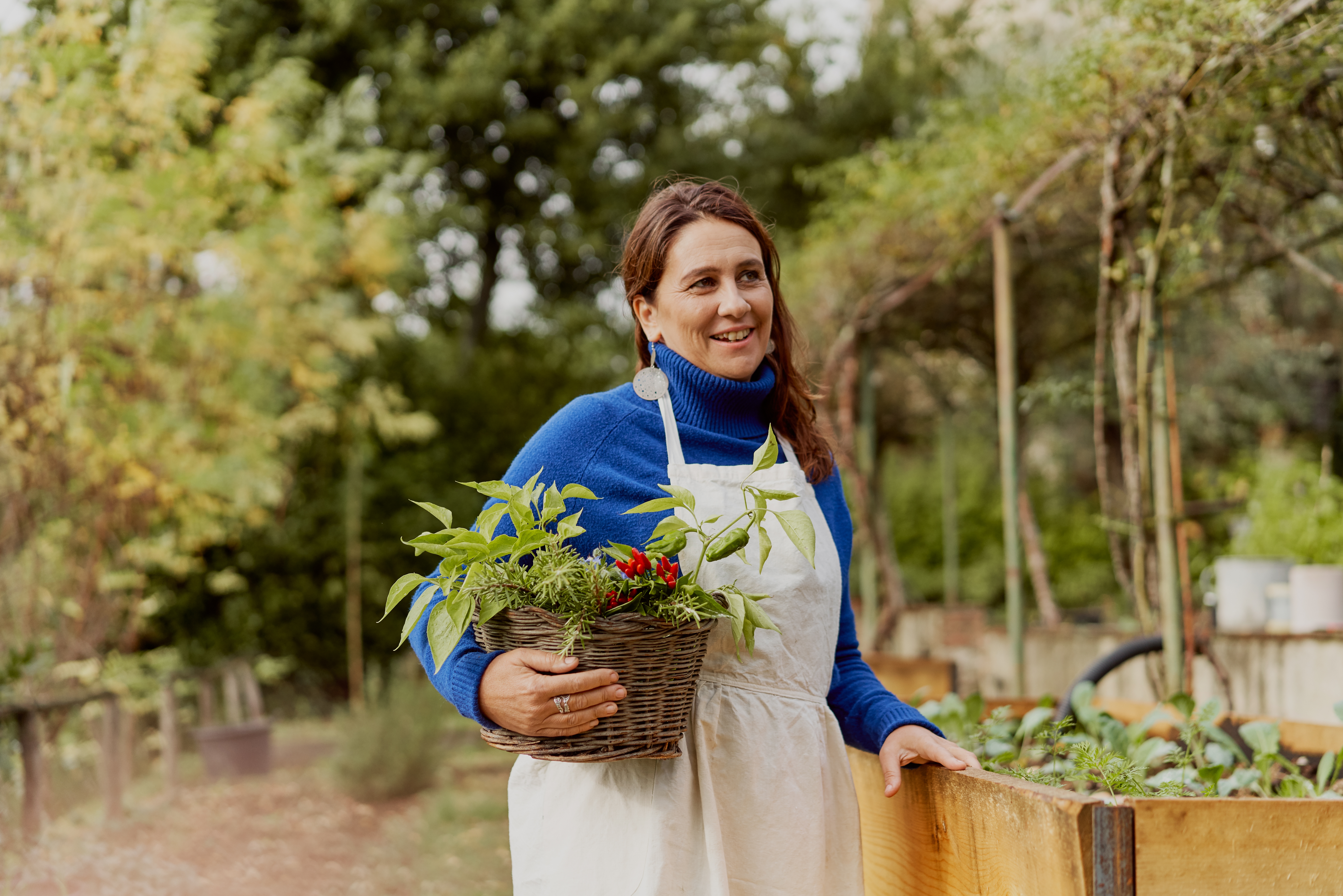 Eine Frau mit Schürze hält draußen in einem Garten einen Korb mit Pflanzen.