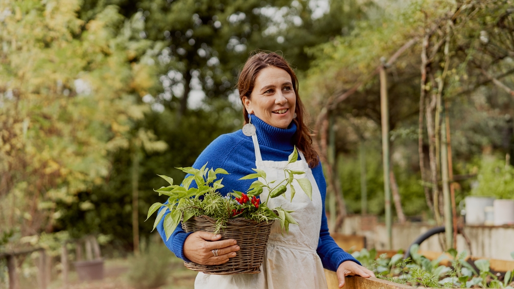 A woman in an apron holds a basket of plants outside in a garden.