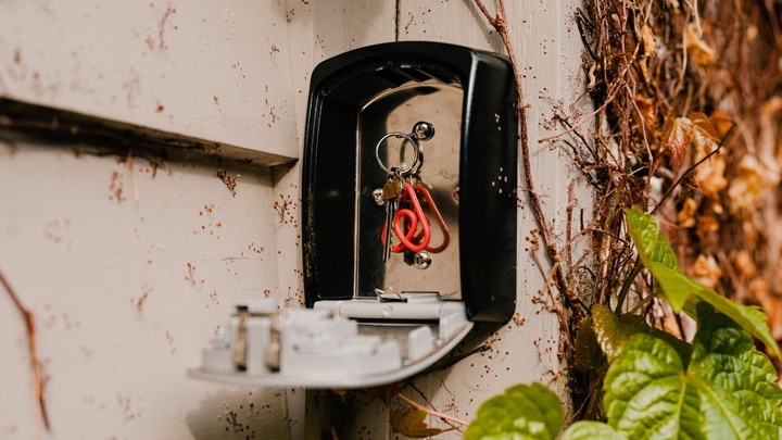 A red Airbnb logo keychain holding a key hangs inside an open metal lockbox that’s on a wall with ivy growing on it.
