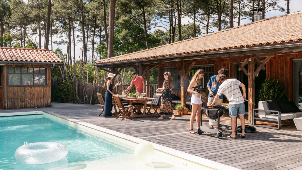 Three people set an outdoor table while three others cook food on a grill that’s situated on a deck next to a swimming pool.