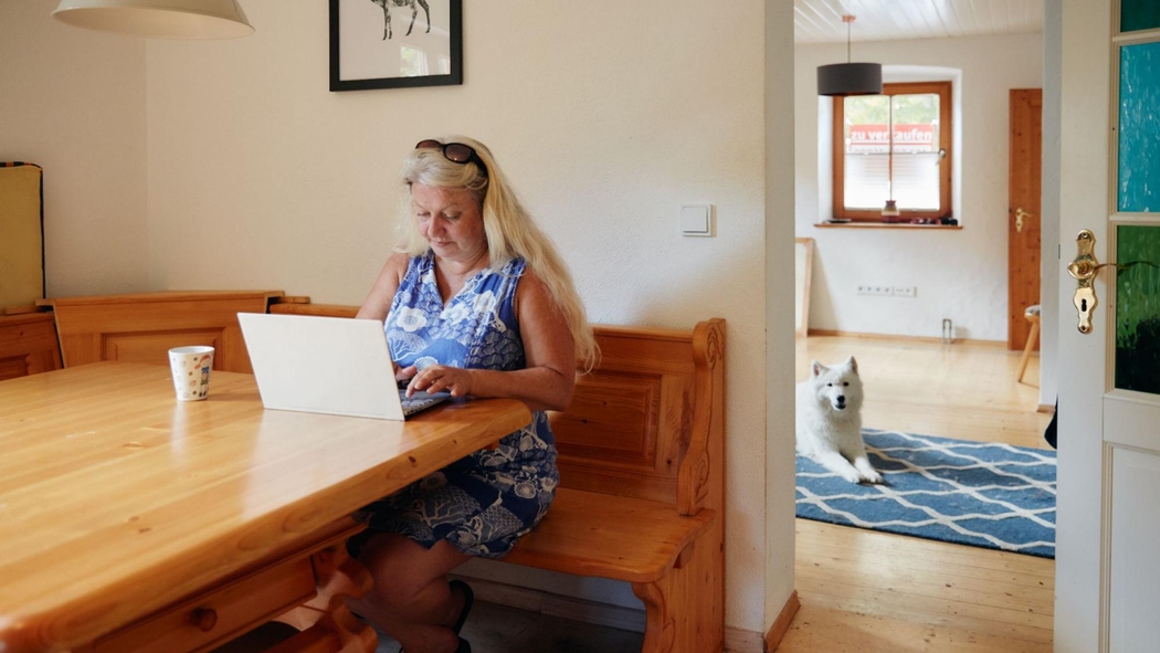 Een persoon zit aan tafel en typt op een laptop terwijl een grote witte hond door de deuropening van een aangrenzende kamer kijkt.