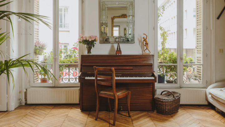 Full-length windows on either side of an upright piano look past a balcony with flowers onto a courtyard below in this Airbnb stay.