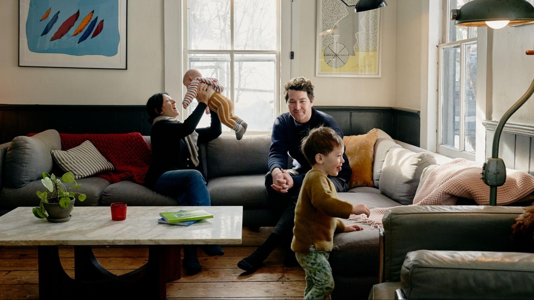 Dos adultos están sentados en un sofá. Uno le sonríe a un bebé que está sosteniendo en el aire mientras el otro observa a un niño pequeño caminar.