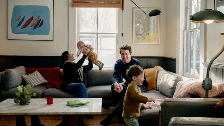 Kaks täiskasvanut istuvad diivanil. Üks hoiab imikut õhus ja naeratab ning teine vaatab mudilast kõndimas.