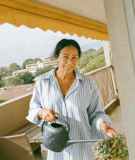 A person holding a watering can on a deck smiles, with trees and sky in the background.