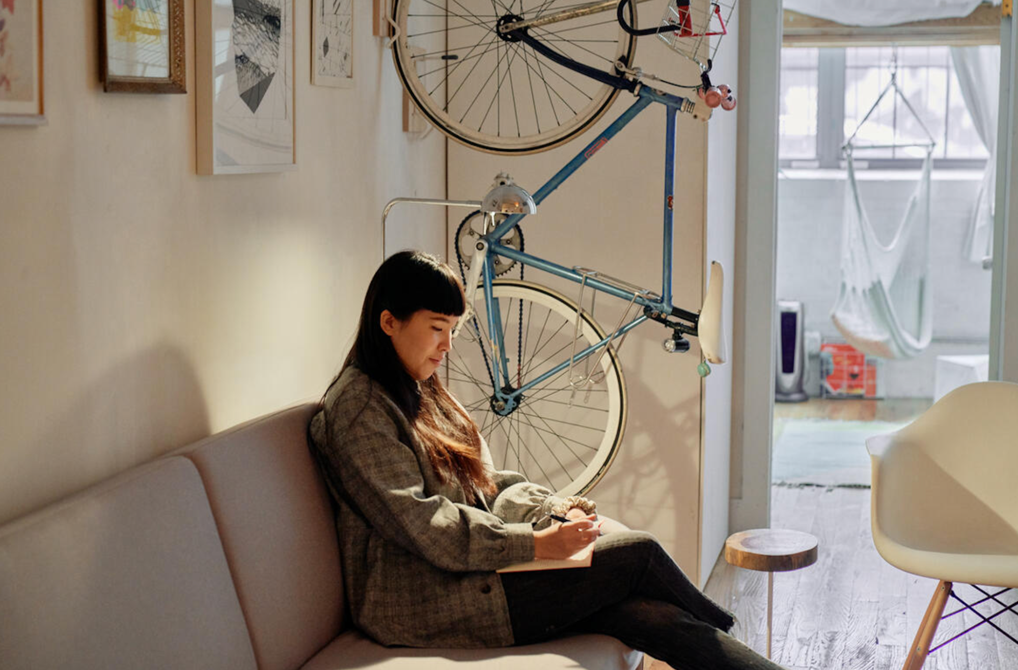 Una persona con el pelo largo está sentada en un sofá y escribe en un diario. Detrás de ella hay una bicicleta y cuadros colgados en la pared.
