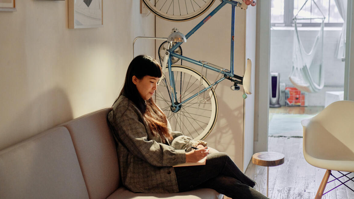 Une femme aux cheveux longs assise sur un canapé écrit dans un journal avec un stylo. Un vélo et plusieurs œuvres d'art encadrées sont accrochés au mur derrière elle, et un hamac est suspendu au plafond dans la pièce voisine.