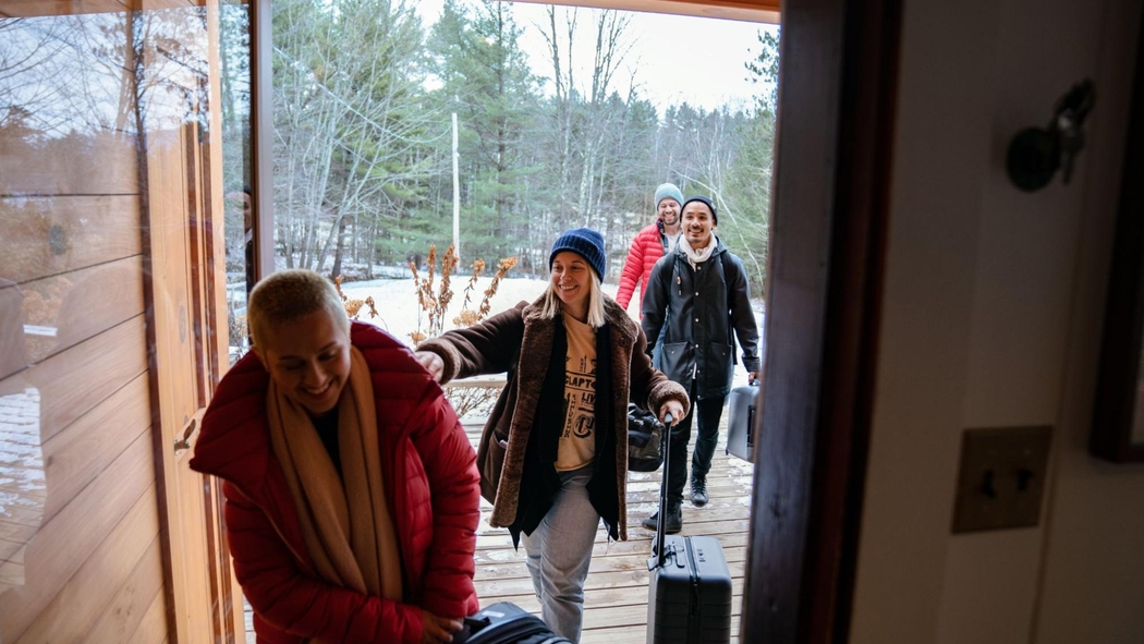 Cuatro personas sonrientes con maletas caminan a través de una puerta principal de vidrio hacia una casa.