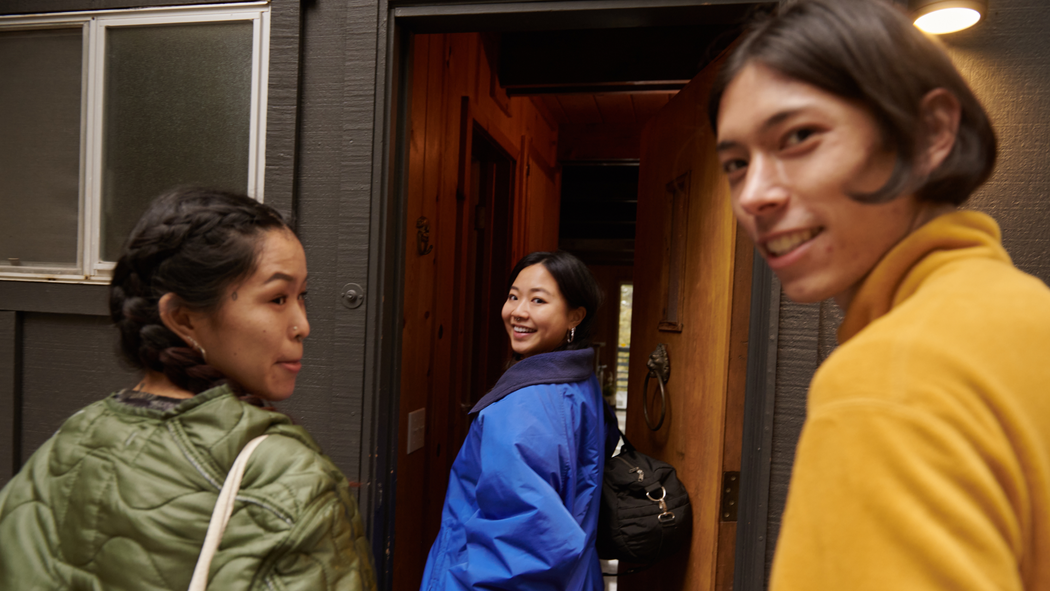 Drei lächelnde Personen, die unterschiedliche Taschen tragen, betreten eine Unterkunft. Die Tür hat ein Fenster über einem bronzenen Türklopfer in Form eines Löwens.