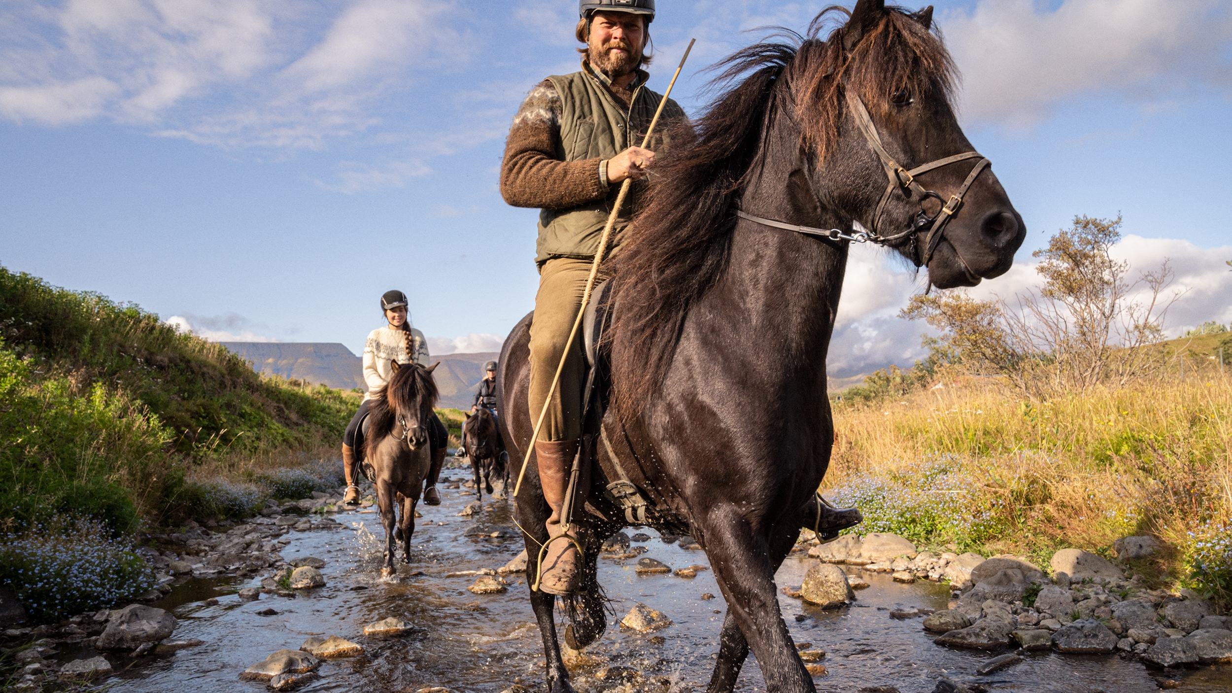 Kask ve kazak giymiş üç kişi, çimenli yamaçlarla çevrili sığ bir dereden atlarıyla geçiyor.