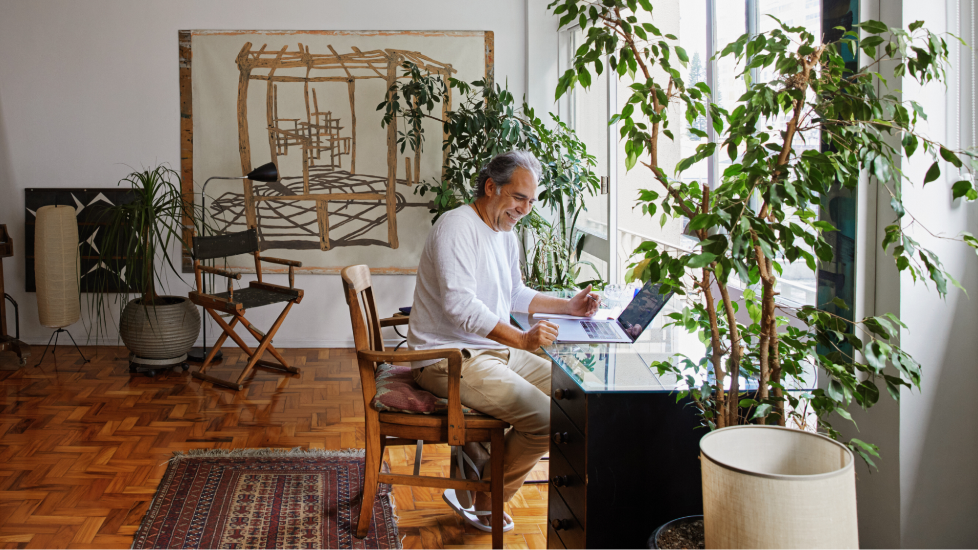 คนนั่งอยู่ที่โต๊ะกระจก มีแล็ปท็อปตั้งอยู่ด้านหน้า แสงอาทิตย์ส่องลงบนพื้นปาร์เก้และกระถางต้นไม้ขนาดใหญ่