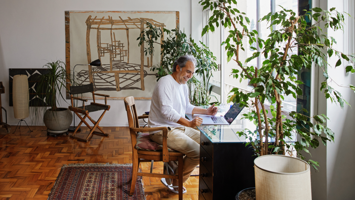 Une personne est assise devant un ordinateur portable posé sur un bureau en verre. Le soleil éclaire le parquet et une grande plante en pot.