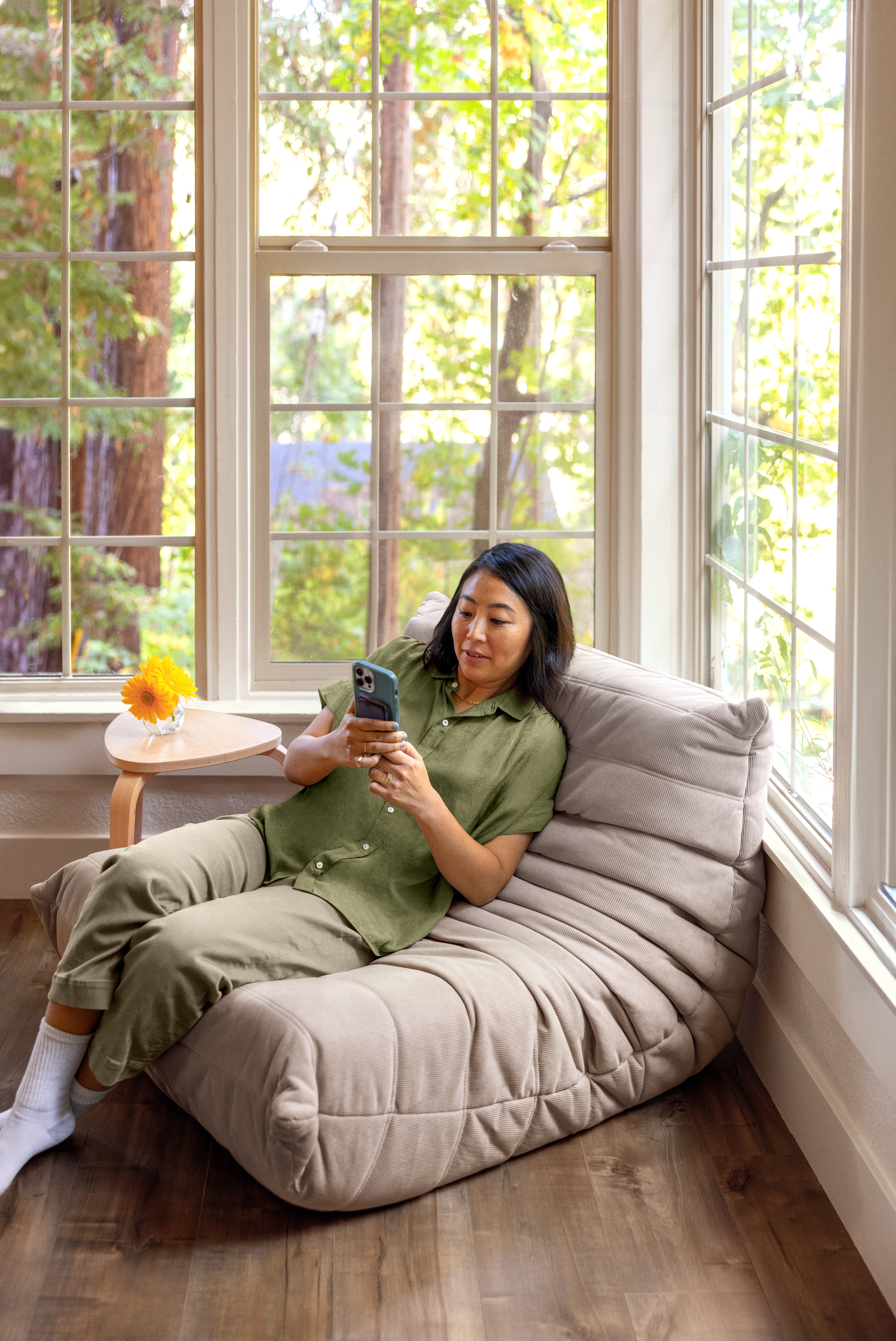 En person i en olivengrønn skjorte med knapper ser på en smarttelefon og lener seg tilbake i en beige stol ved siden av vinduer med utsikt over trær.