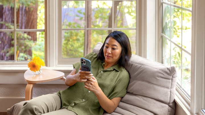 Una persona con una camisa verde mira un smartphone sentada en una silla beige al lado de una ventana con vistas a unos árboles.