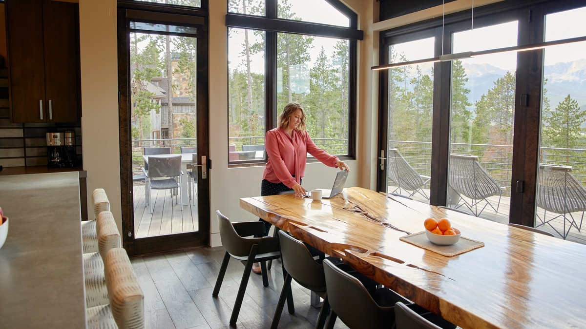 Une personne se tient à l'extrémité d'une longue table à manger en bois, devant un ordinateur portable ouvert. La pièce dispose de baies vitrées.