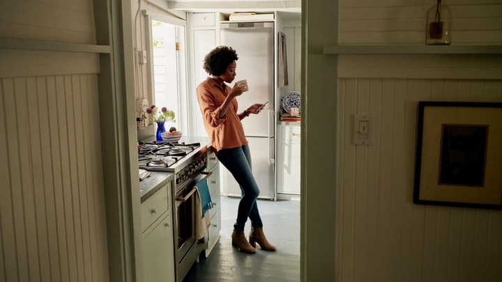 A person wearing a button-up shirt leans against a kitchen countertop while holding a mug and looking at a mobile phone.