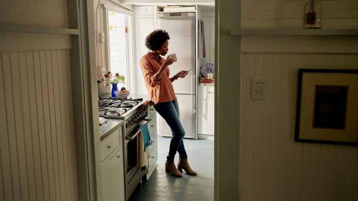 Eine Person, die ein Hemd mit Knopfleiste trägt, lehnt sich an eine Küchenarbeitsplatte, während sie eine Tasse in der Hand hält und auf ein Handy schaut.
